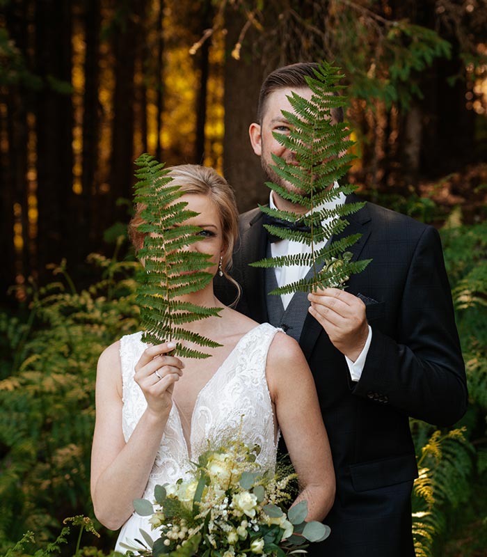 Verliebtes Brautpaar versteckt sein Gesicht hinter einem Farnblatt während eines romantischen Waldspaziergangs in Karlsruhe. Hochzeitsfotografie von Wagner Photography.